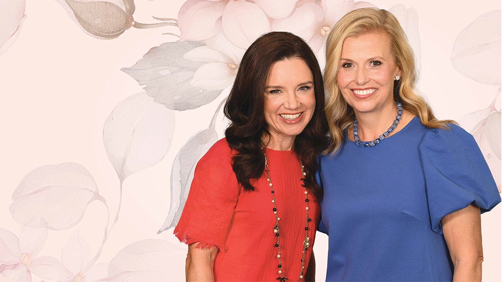 Two smiling women standing together against a soft floral background, dressed in red and blue outfits for a Dallas event.