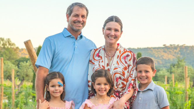 Smiling family of five in a vineyard, with parents standing behind their three children on a sunny day.