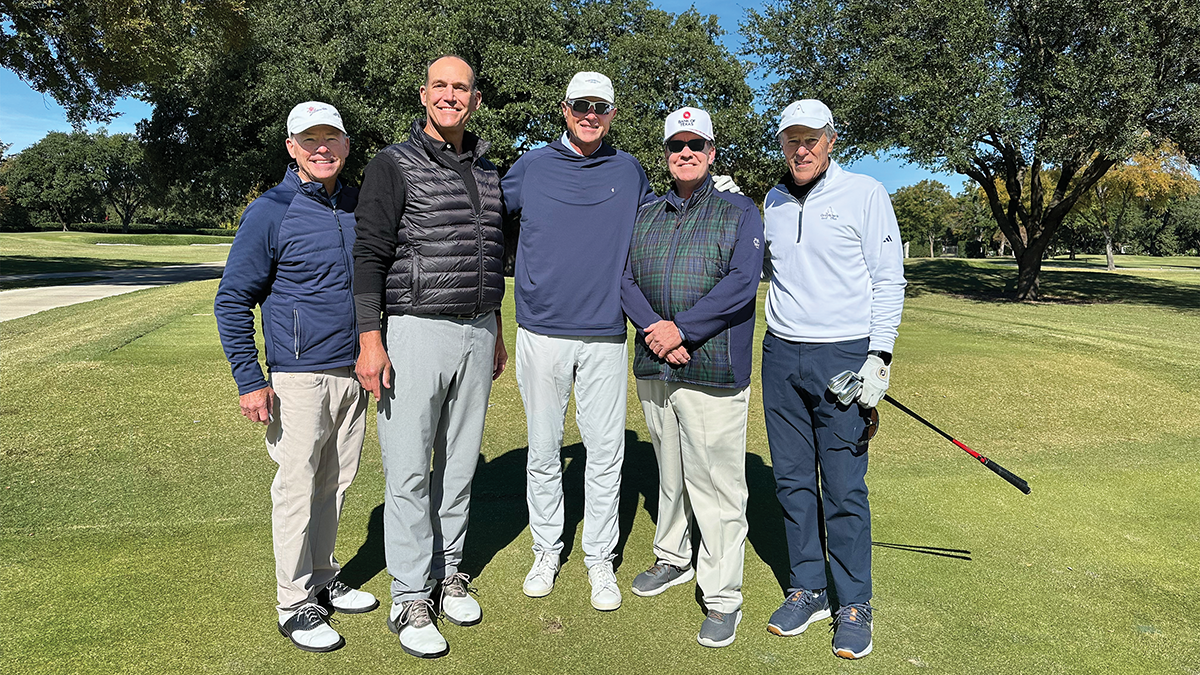 Five men smiling together on a golf course on a sunny day, surrounded by trees.