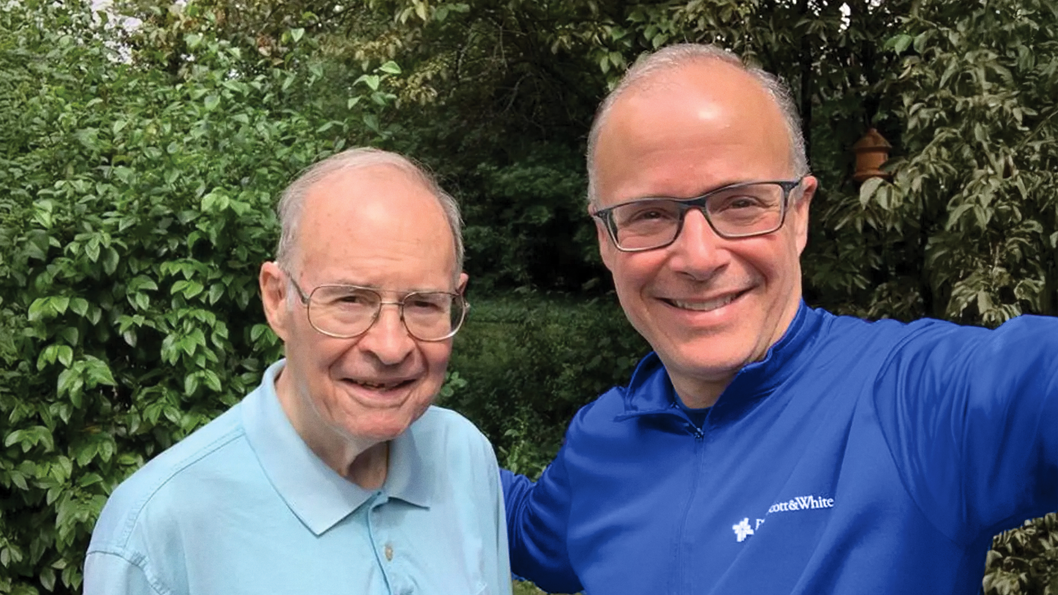 Two men smiling outdoors, standing together in a garden setting, with one wearing a Baylor Scott & White jacket.